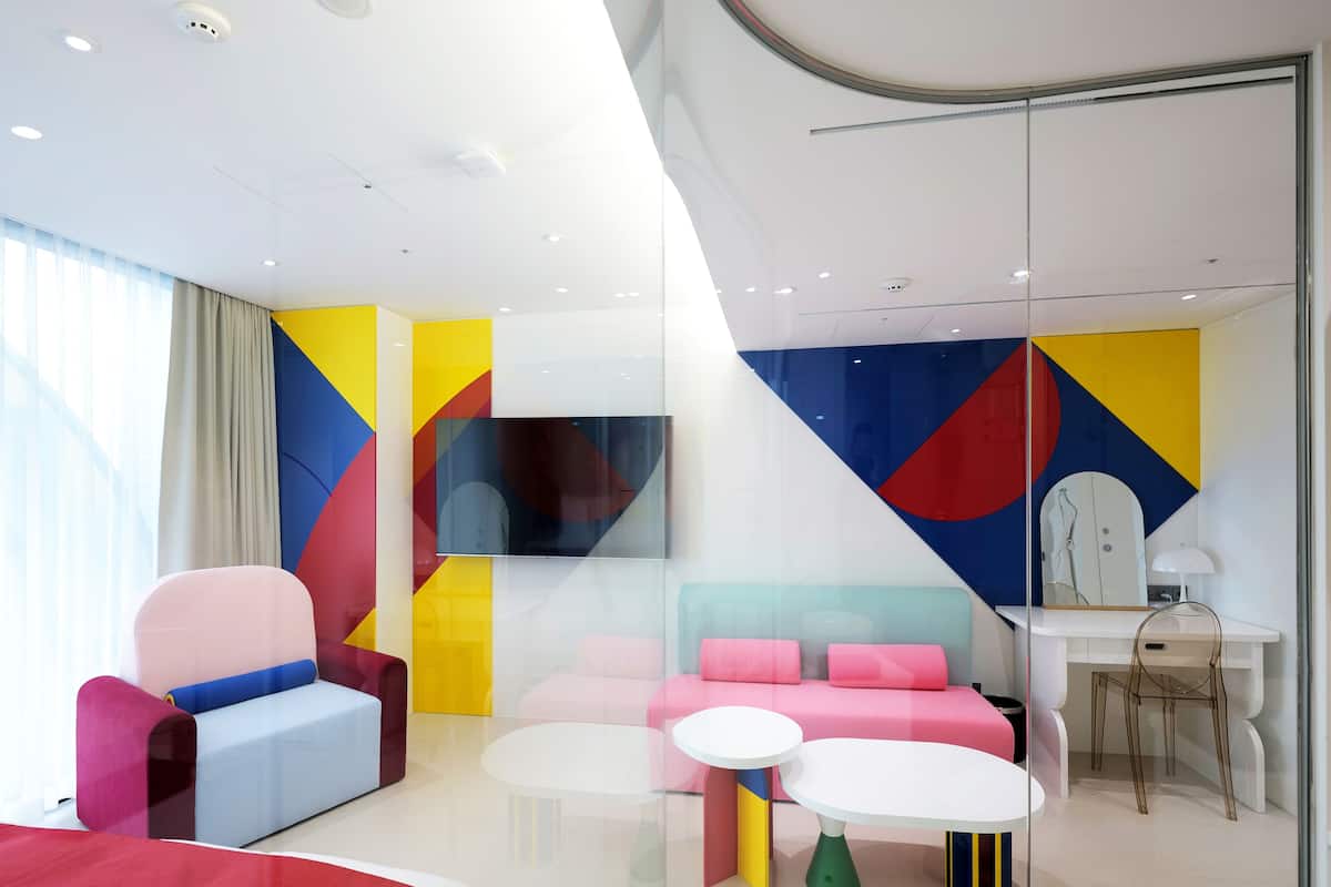 Une chambre d’hôtel moderne avec des œuvres d’art géométriques colorées sur les murs, un téléviseur à écran plat, un bureau avec une lampe et un coin salon comprenant un canapé et des fauteuils.