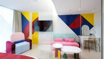 A modern hotel room with colorful geometric wall art, a flat-screen TV, a desk with a lamp, and a seating area with a sofa and chairs.