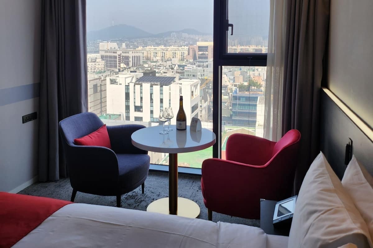 Une chambre d’hôtel avec un lit, deux chaises, une petite table avec une bouteille et des verres, et une grande fenêtre donnant sur la ville.