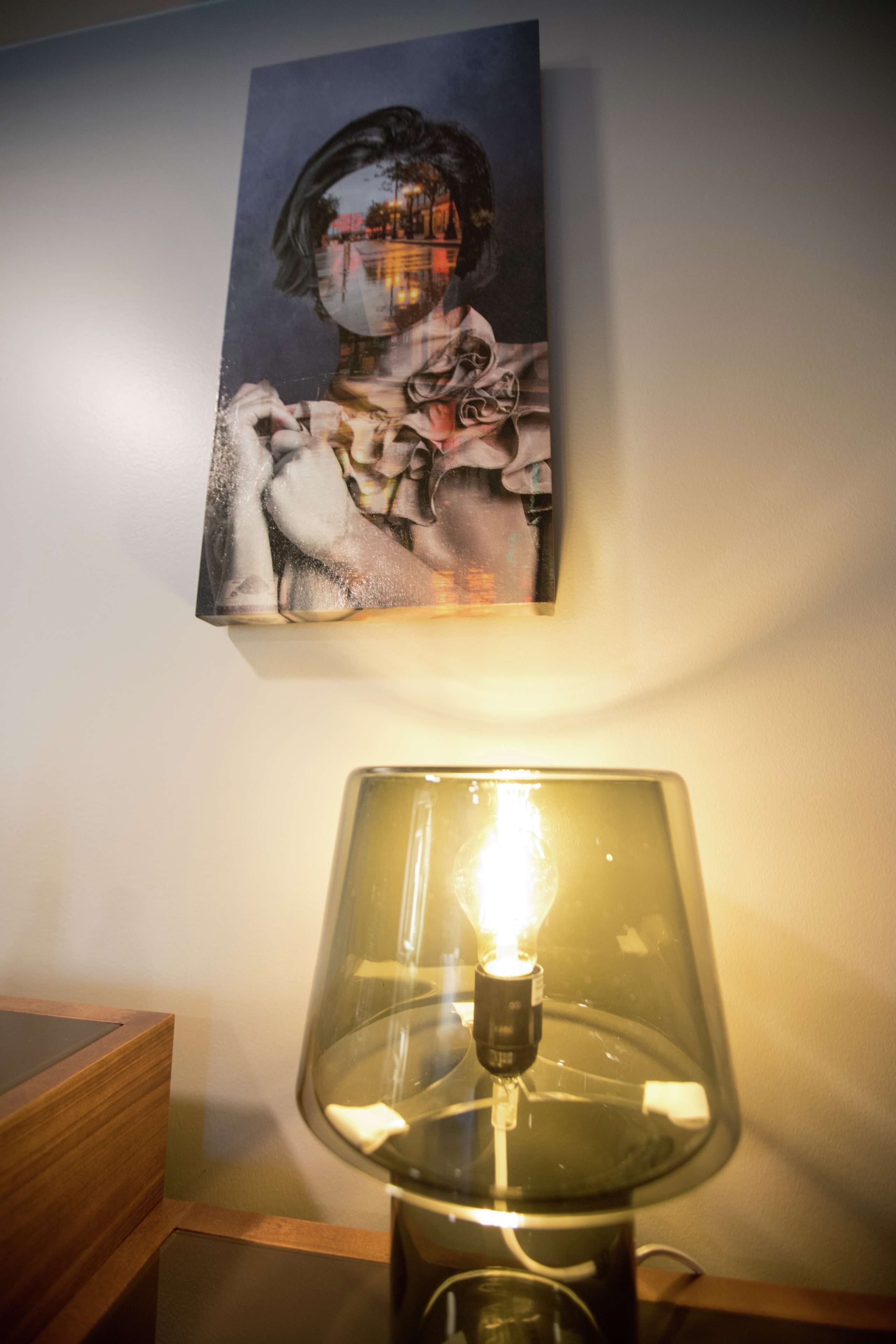 A wall-mounted painting of a person with a blurred face, a table lamp with a visible light bulb, and a wooden side table.