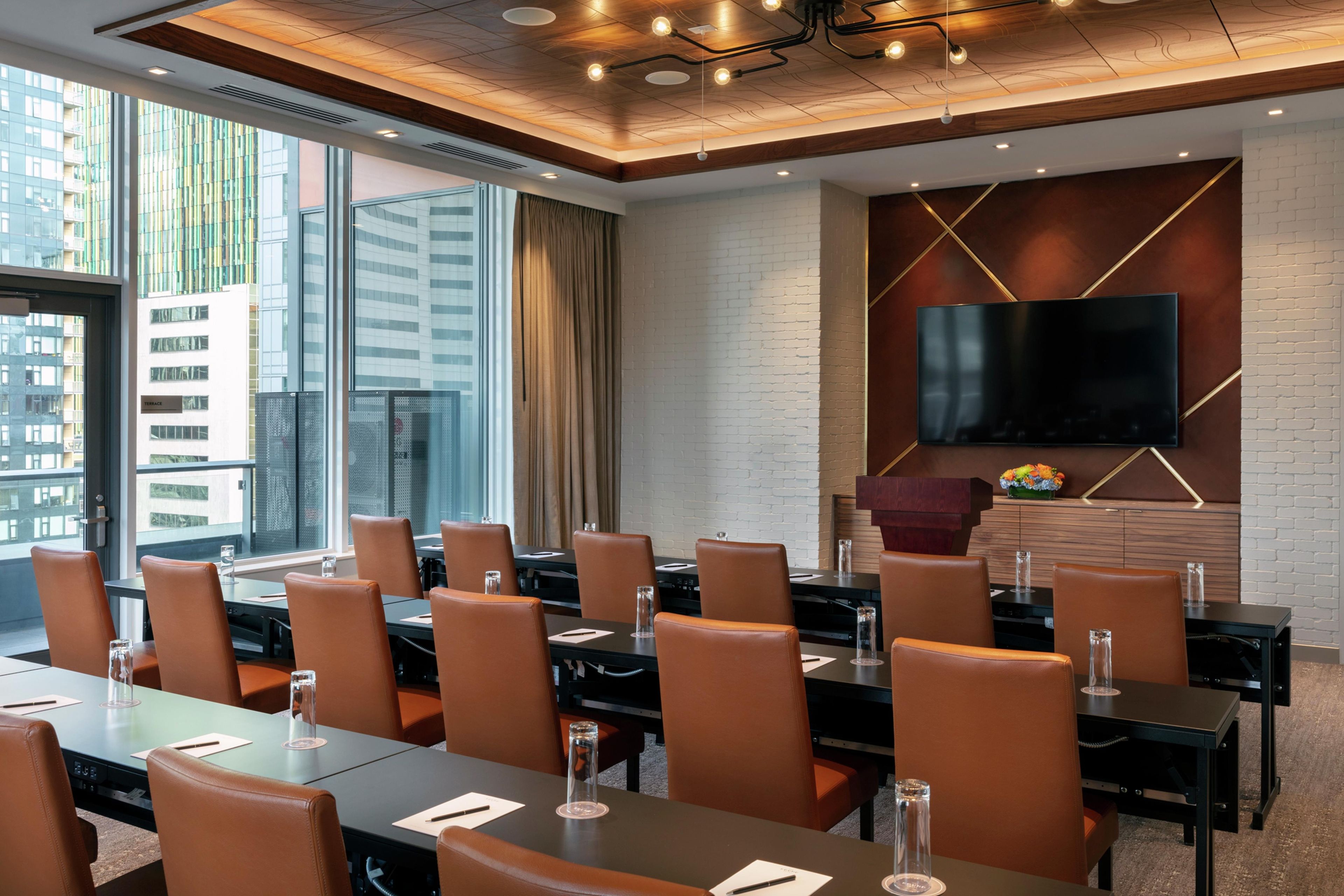 A conference room with a large flat-screen TV mounted on a brick wall, leather chairs, and tables with water glasses.