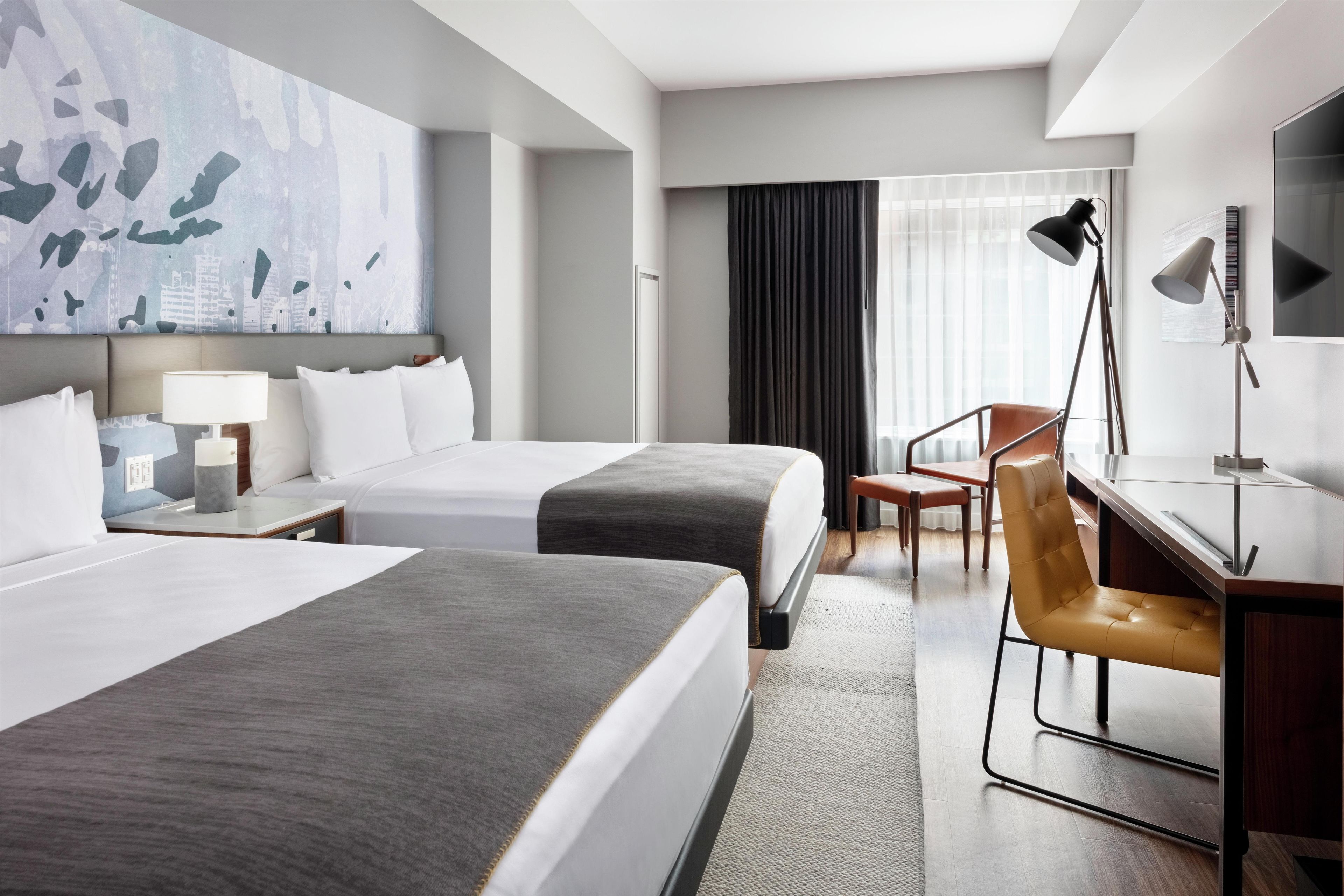 Habitación de hotel con dos camas, un escritorio, una silla, una lámpara y un cuadro abstracto grande en la pared.