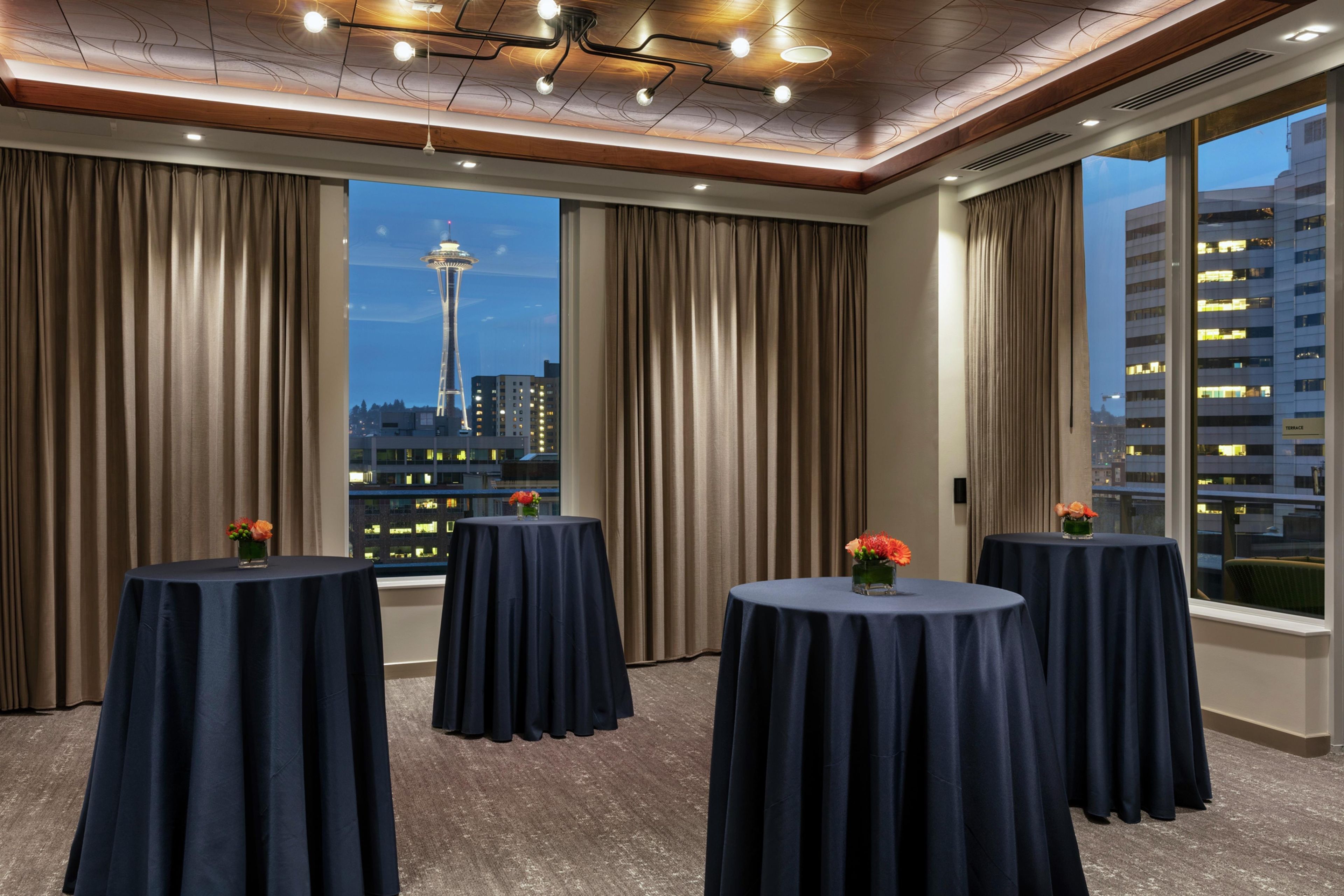 Habitación de hotel con cuatro mesas redondas, ventanas amplias con cortinas y vistas a un horizonte urbano, además de un diseño moderno en el techo.