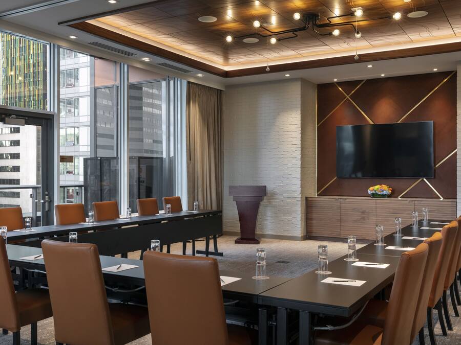 A conference room with a long table, brown chairs, a large screen, and a white brick wall.