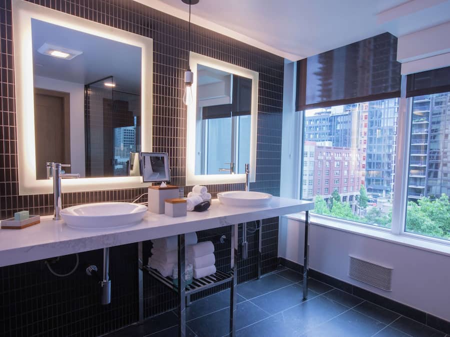 A modern bathroom with two sinks, large mirrors, and a view of a cityscape through a large window.