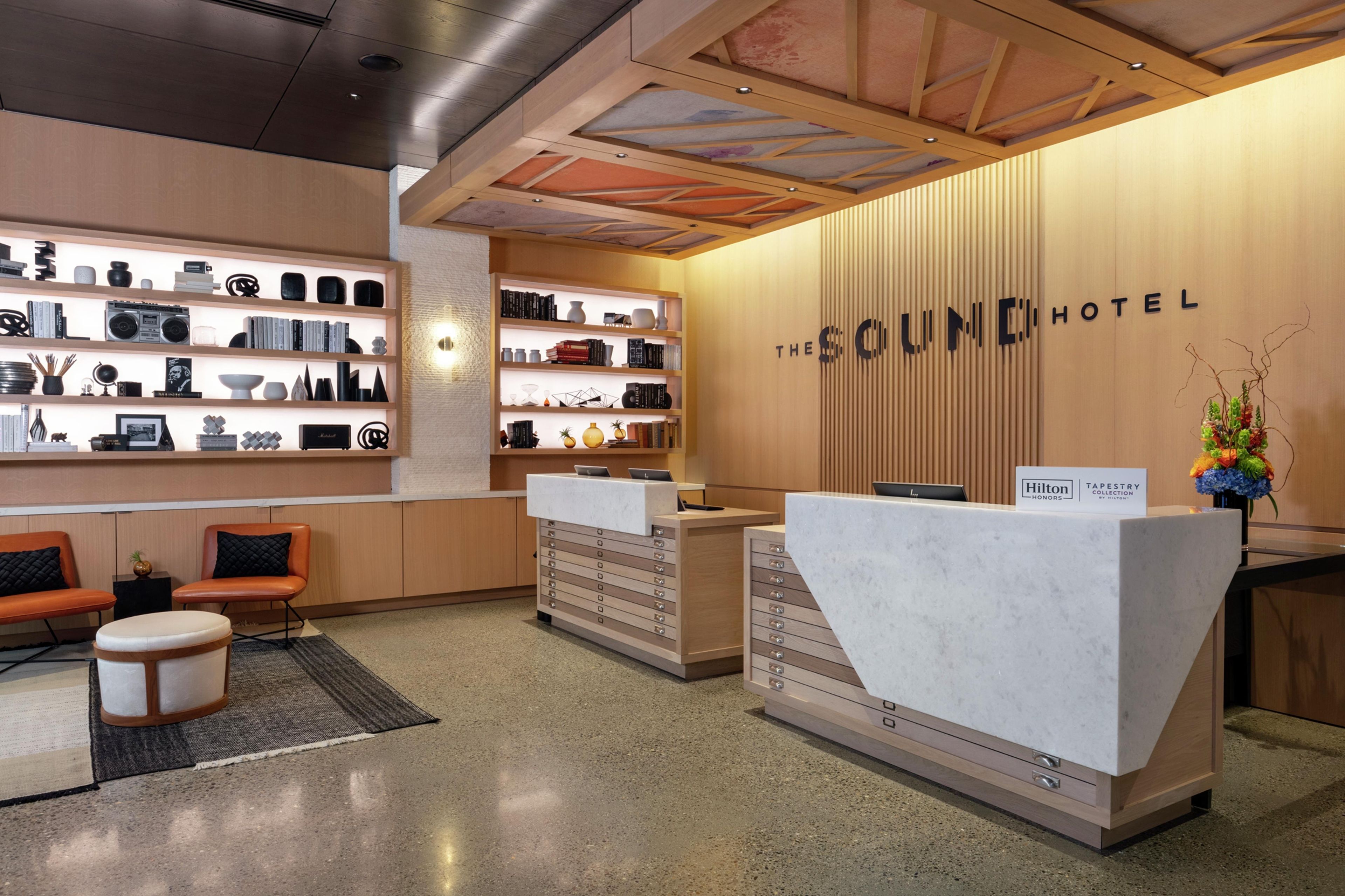 A hotel reception area with a modern design, featuring a reception desk, seating area, and a wall with shelves displaying various items.