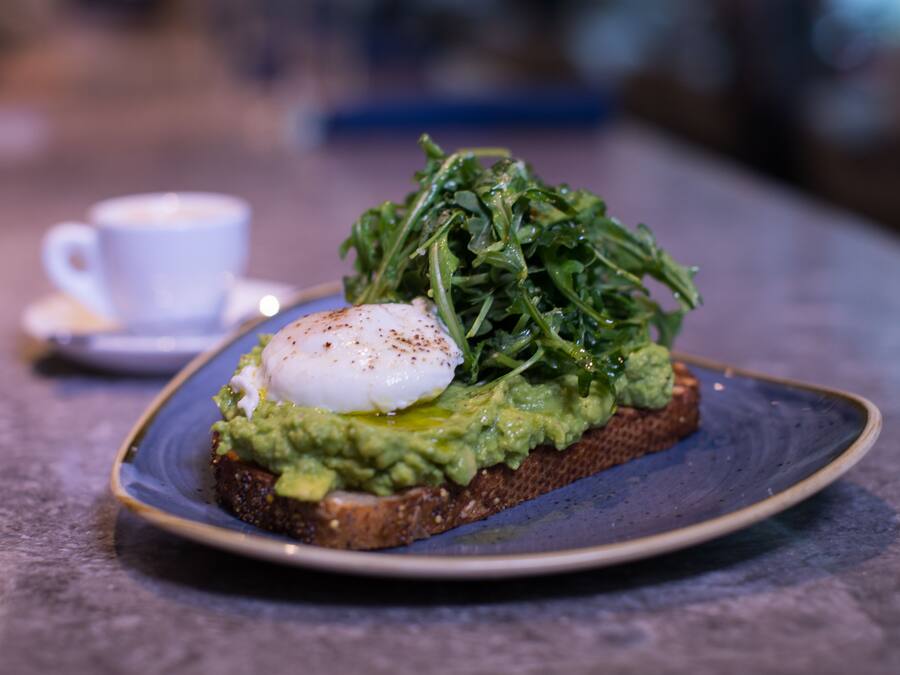A plate with a poached egg, avocado, and greens on a slice of bread, accompanied by a cup of coffee on a saucer.