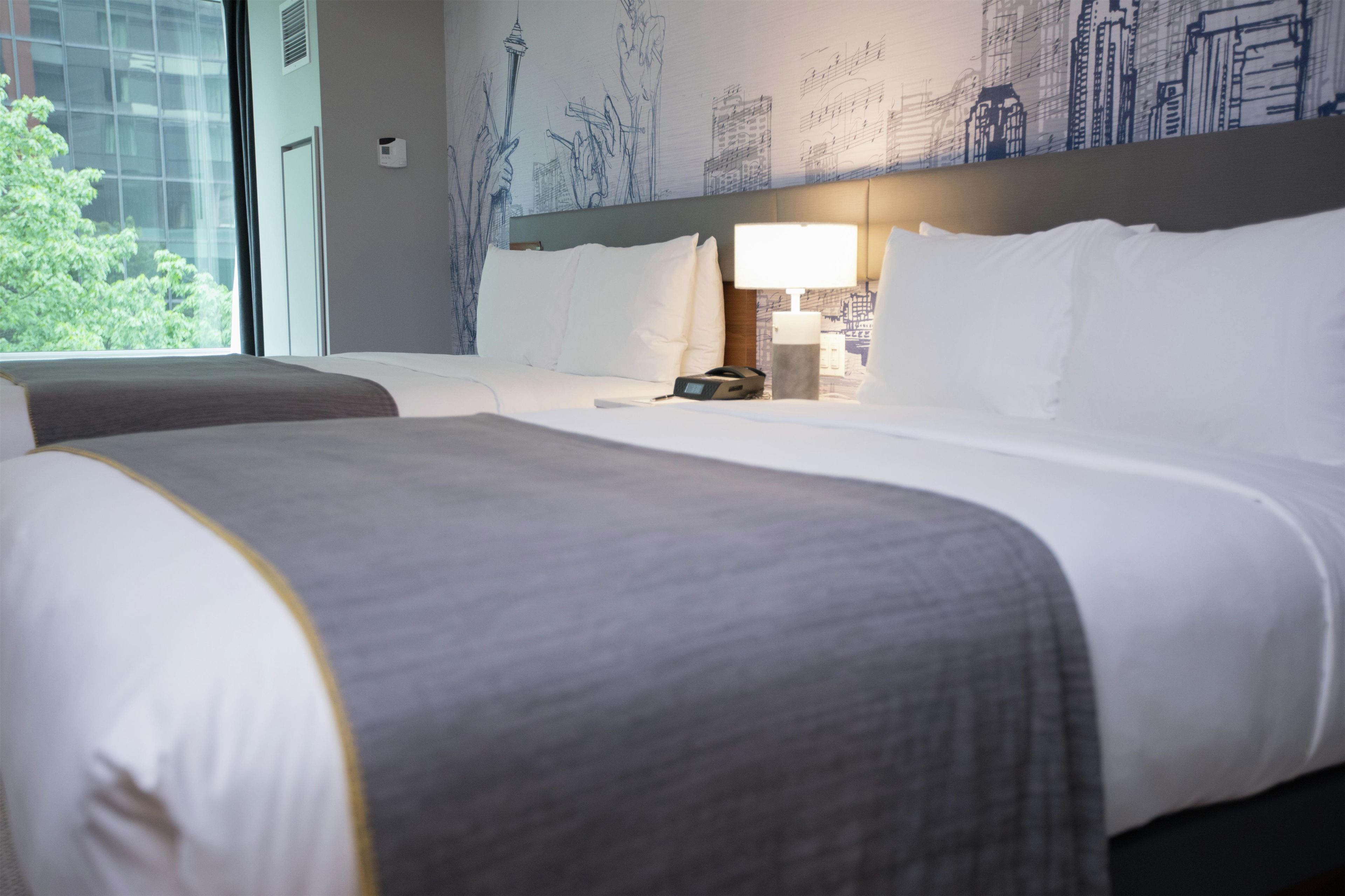 A hotel room with two beds, a cityscape mural on the wall, and a window with a view of trees.