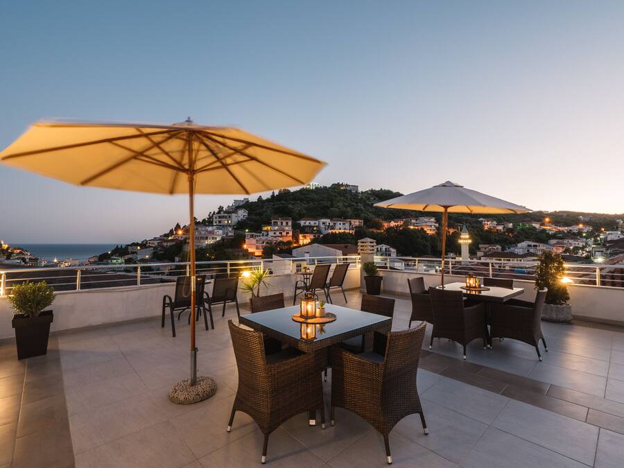 Una terraza en la azotea con una mesa, sillas, un paraguas grande y vistas a un pueblo con edificios y una colina al fondo.