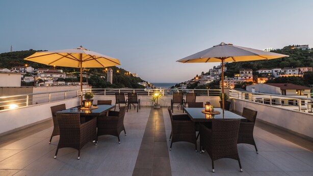Una terraza en la azotea con mesas y sillas para comer, dos sombrillas grandes, y vistas de la ciudad al fondo.