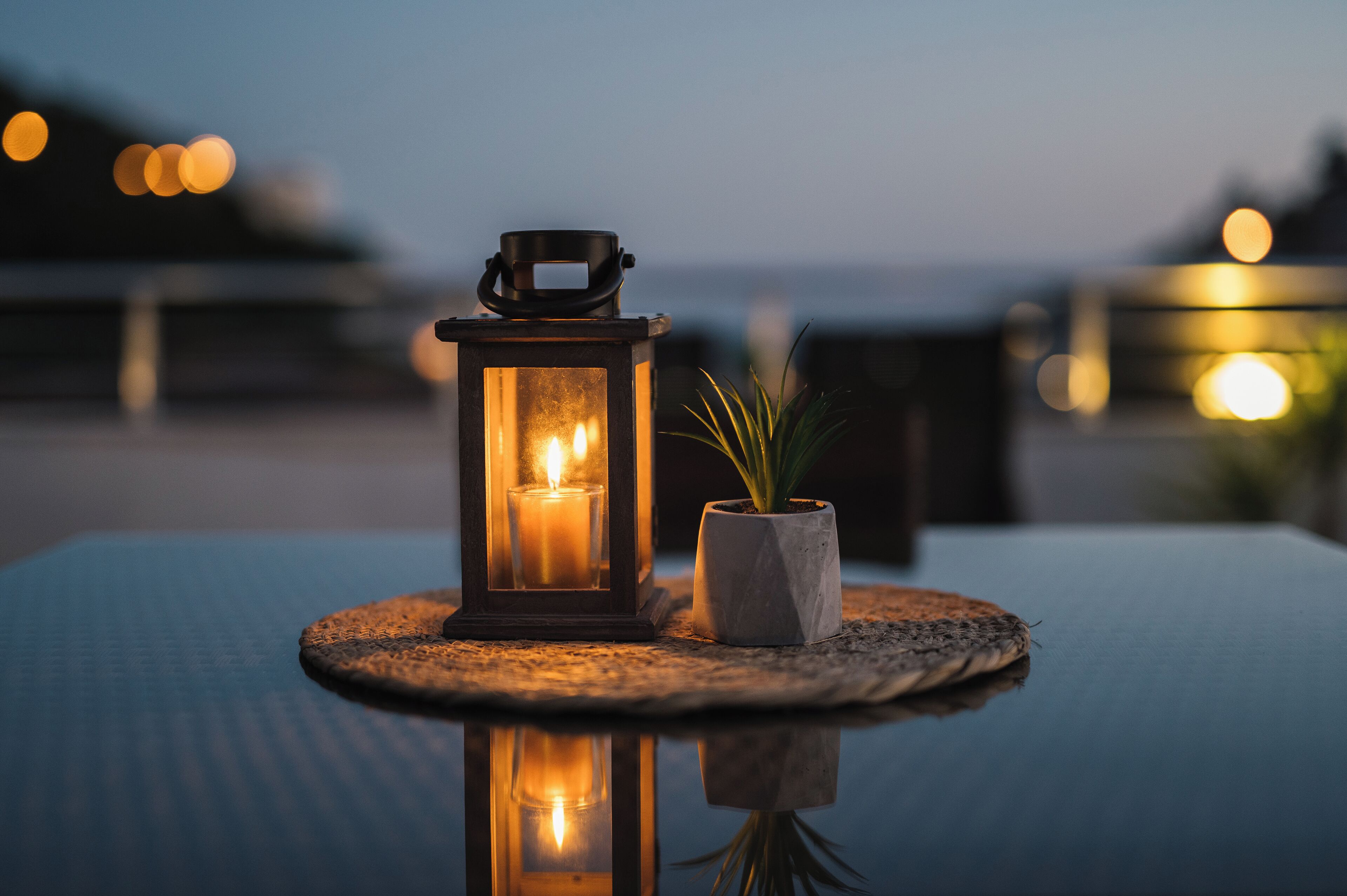 A lantern with a candle on a table, a potted plant, and a view of the sea at dusk.