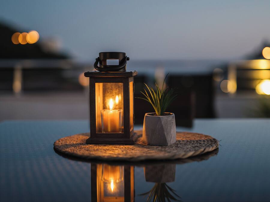 A lantern with a candle on a table, a potted plant, and a view of the sea at dusk.