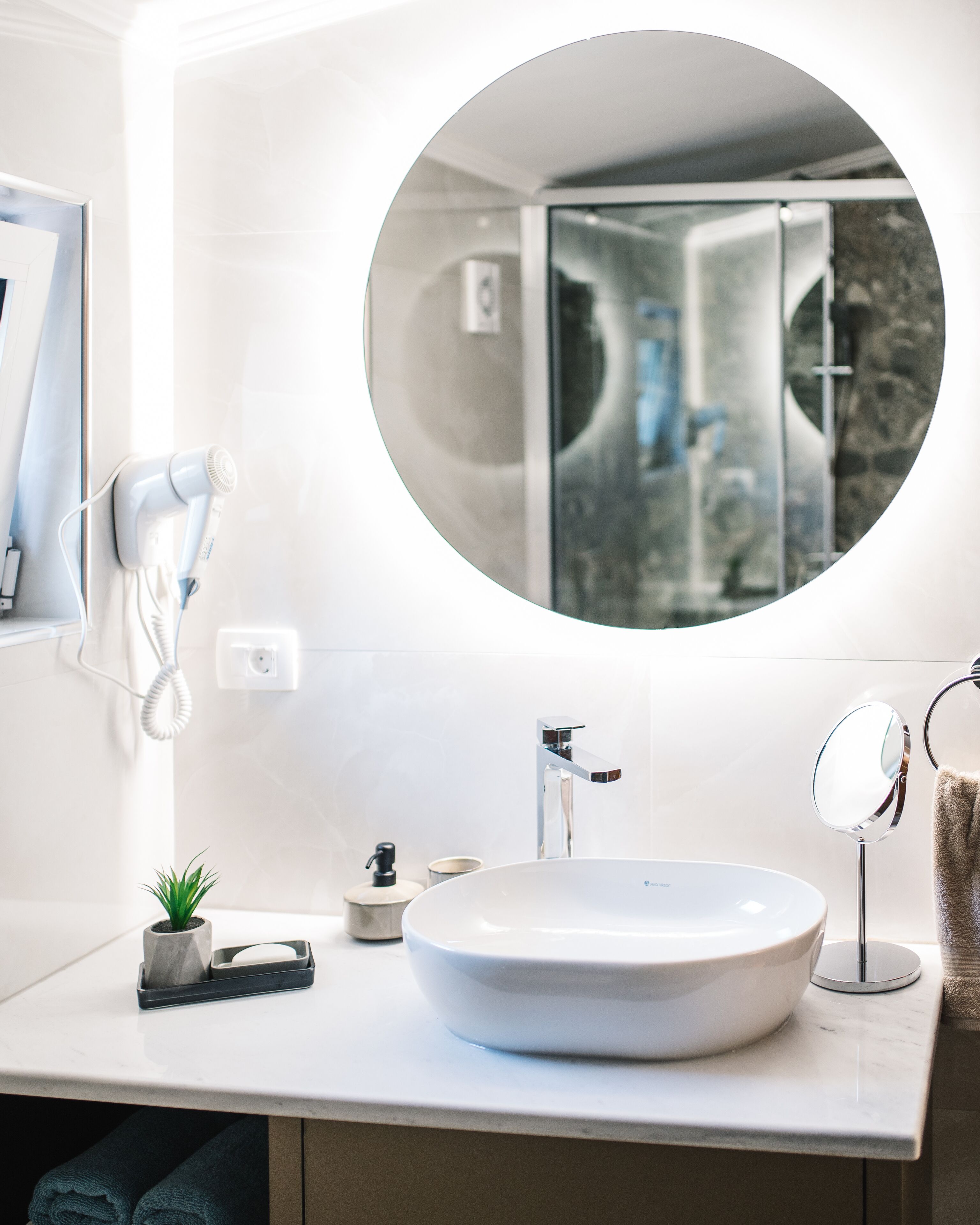 A modern bathroom with a large oval sink, a round mirror, a hairdryer, and a small potted plant.