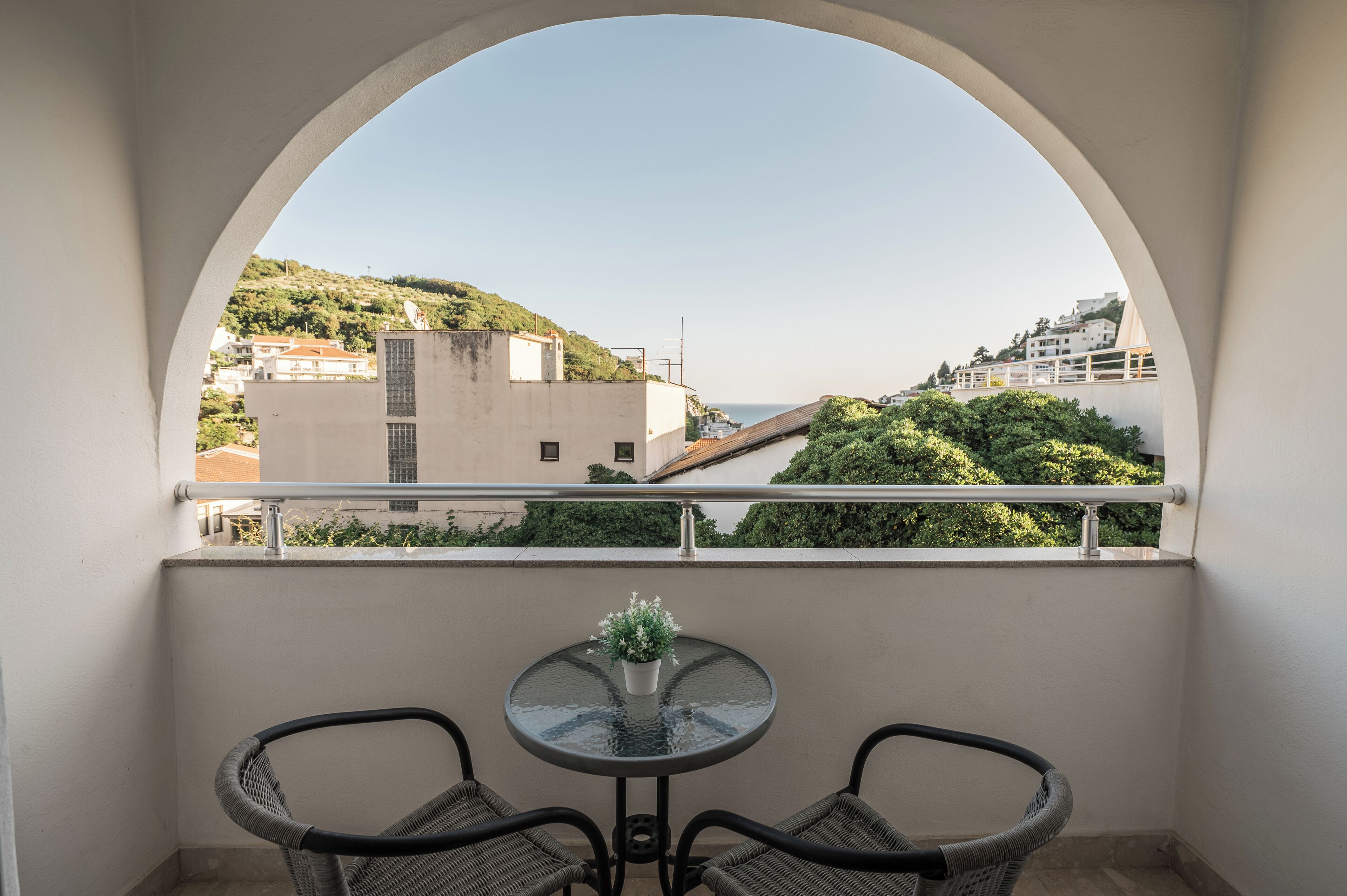 A balcony with a glass table and wicker chairs, overlooking a hillside with buildings and greenery.