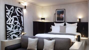 Una habitación de hotel moderna con tragaluces, un cuadro abstracto grande, una cama con ropa blanca, un sofá oscuro y una mesa de centro de vidrio.