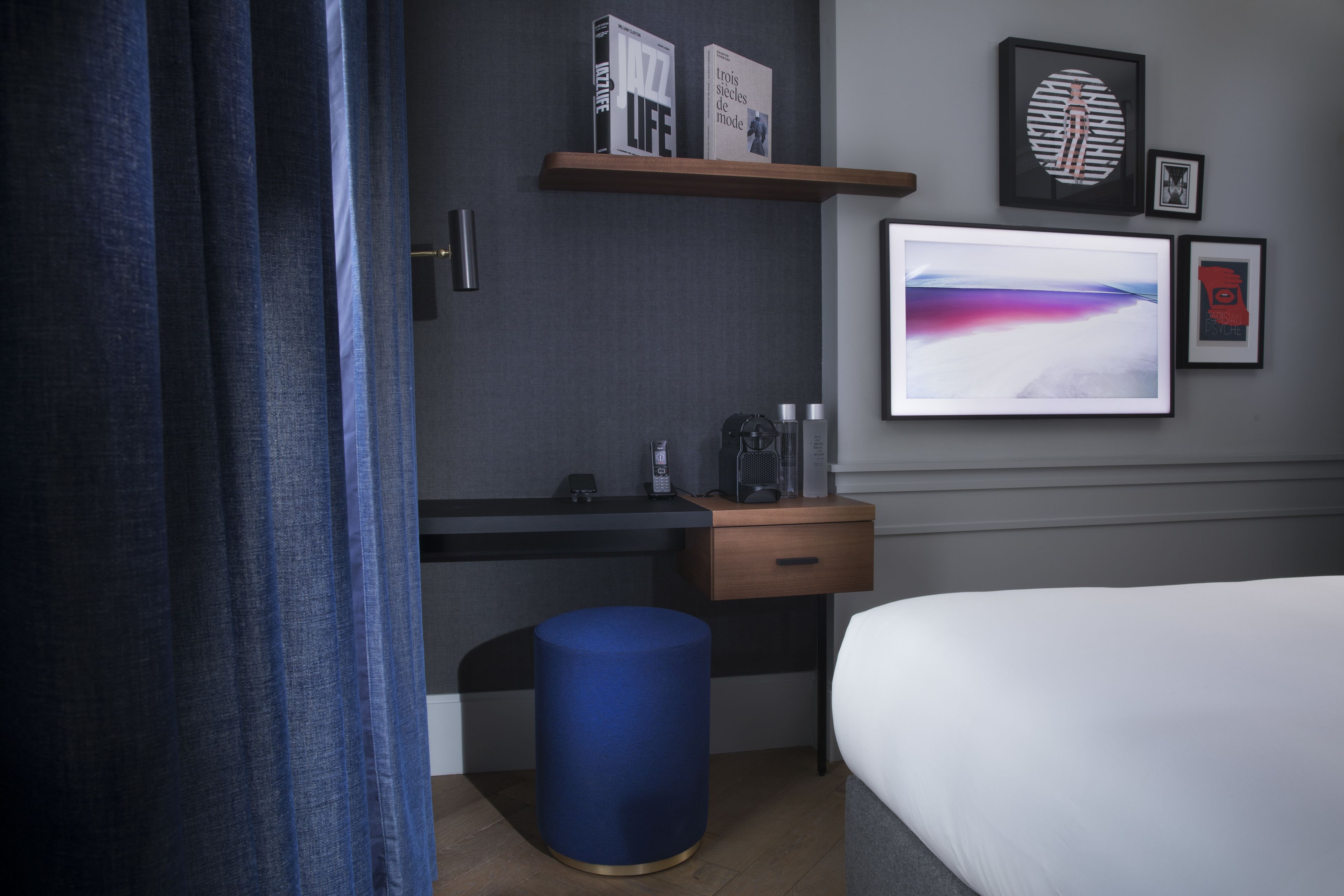 Ein modernes Hotelzimmer mit einem Bett, einem Schreibtisch mit Computer, einem blauen Hocker, einer Kaffeemaschine und gerahmten Kunstwerken an der Wand.