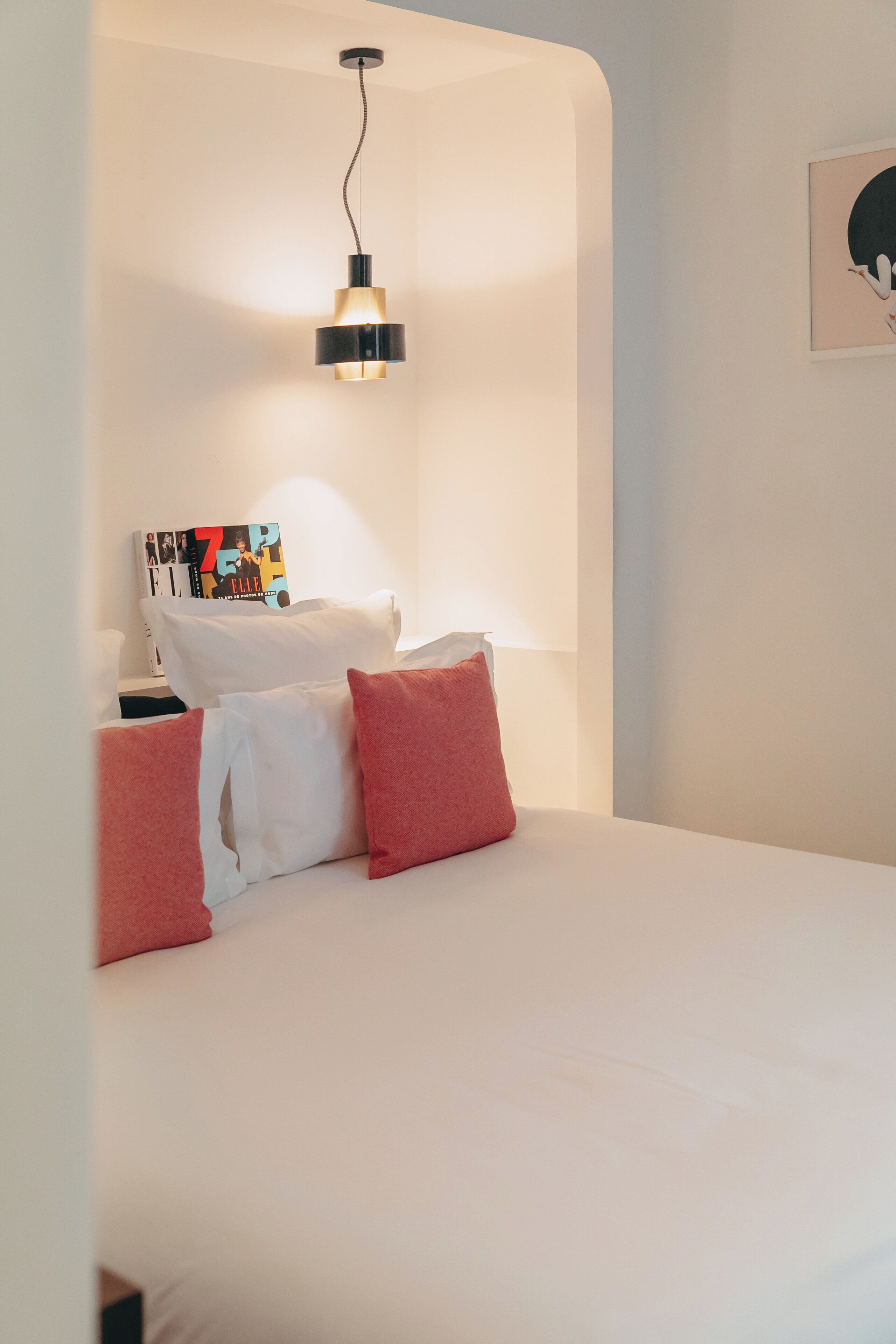 A neatly made bed with white and red pillows, a black hanging lamp, and a framed picture on the wall.