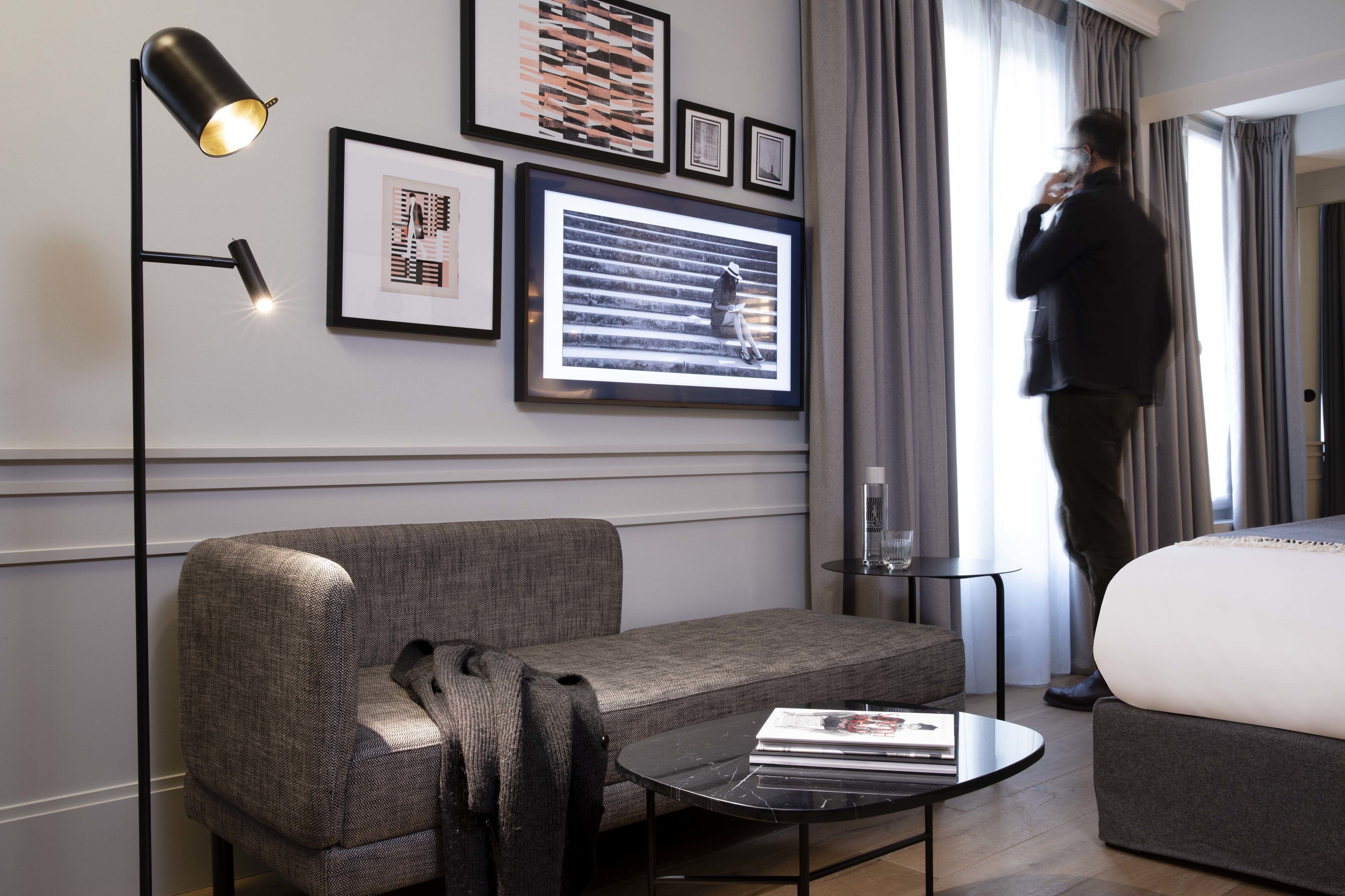 A modern hotel room with a grey sofa, a small round table with a book, a floor lamp, and framed artwork on the wall.