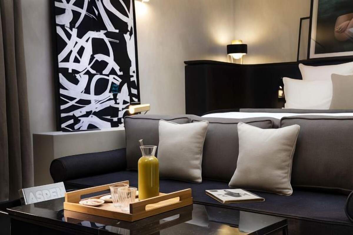 A modern hotel room with a dark gray sofa, a glass coffee table with a tray of cookies and a drink, and a large abstract painting on the wall.