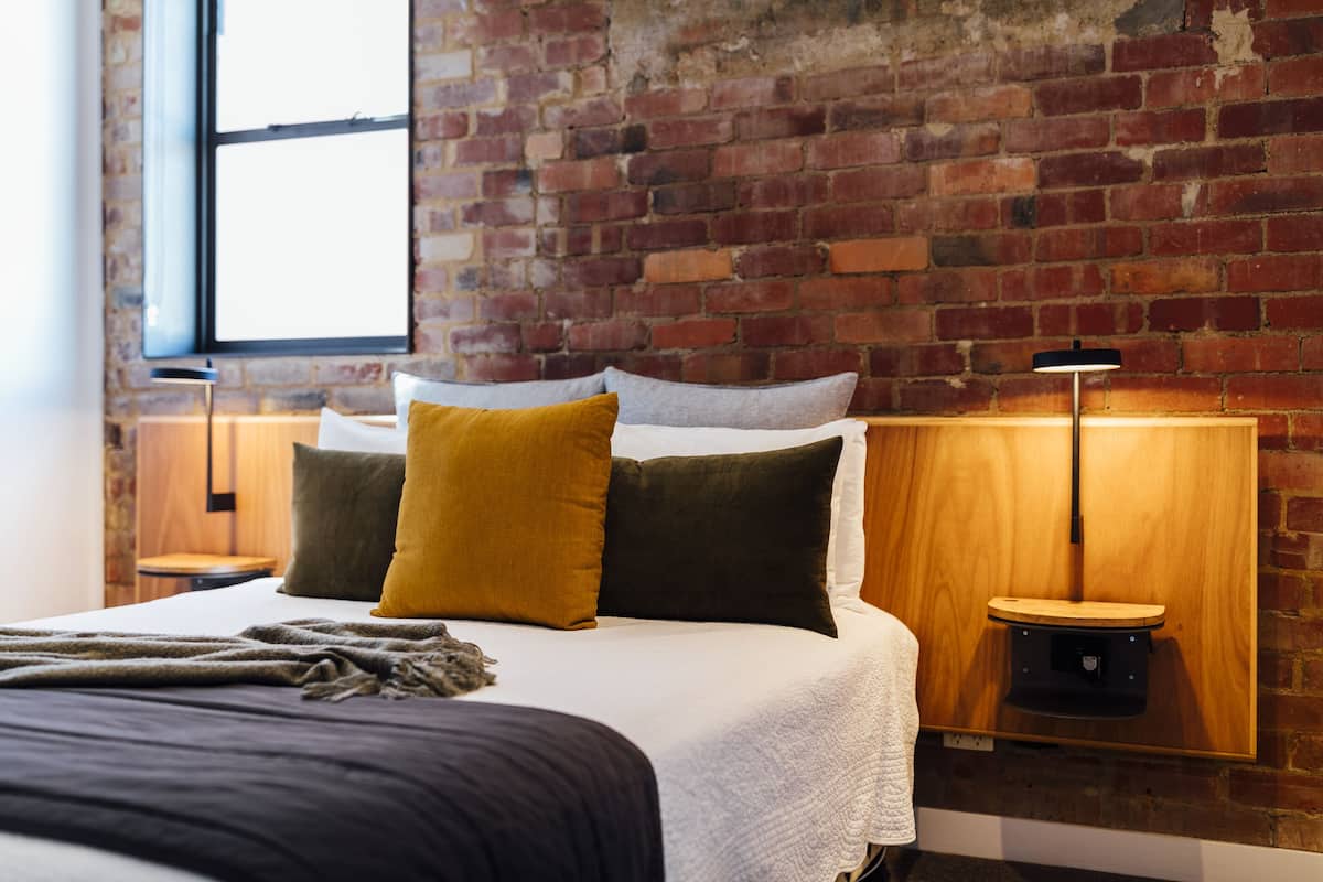 Una habitación de hotel con pared de ladrillo, un ventanal grande, una cama con varias almohadas, una mesita de noche con lámpara y una mesita auxiliar de madera.