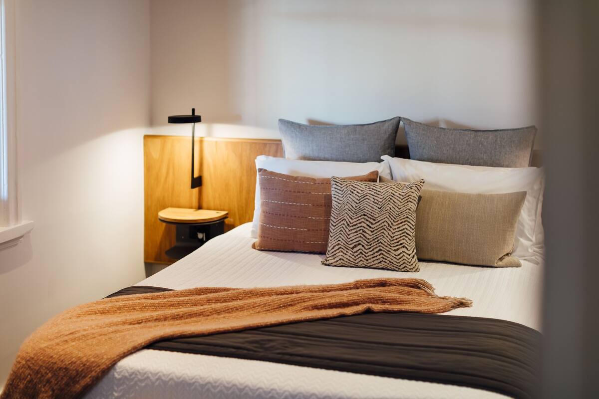 Una cama bien tendida con una manta marrón, una mesita de noche de madera con una lámpara y un taburete de madera pequeño.