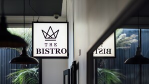 A sign for 'The Bistro' with a crown logo, hanging in a well-lit interior space with large windows and a view of greenery outside.