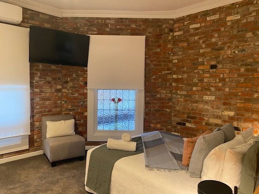 A bedroom with a brick wall, a bed with multiple pillows, a chair, and a window with a view of the outside.