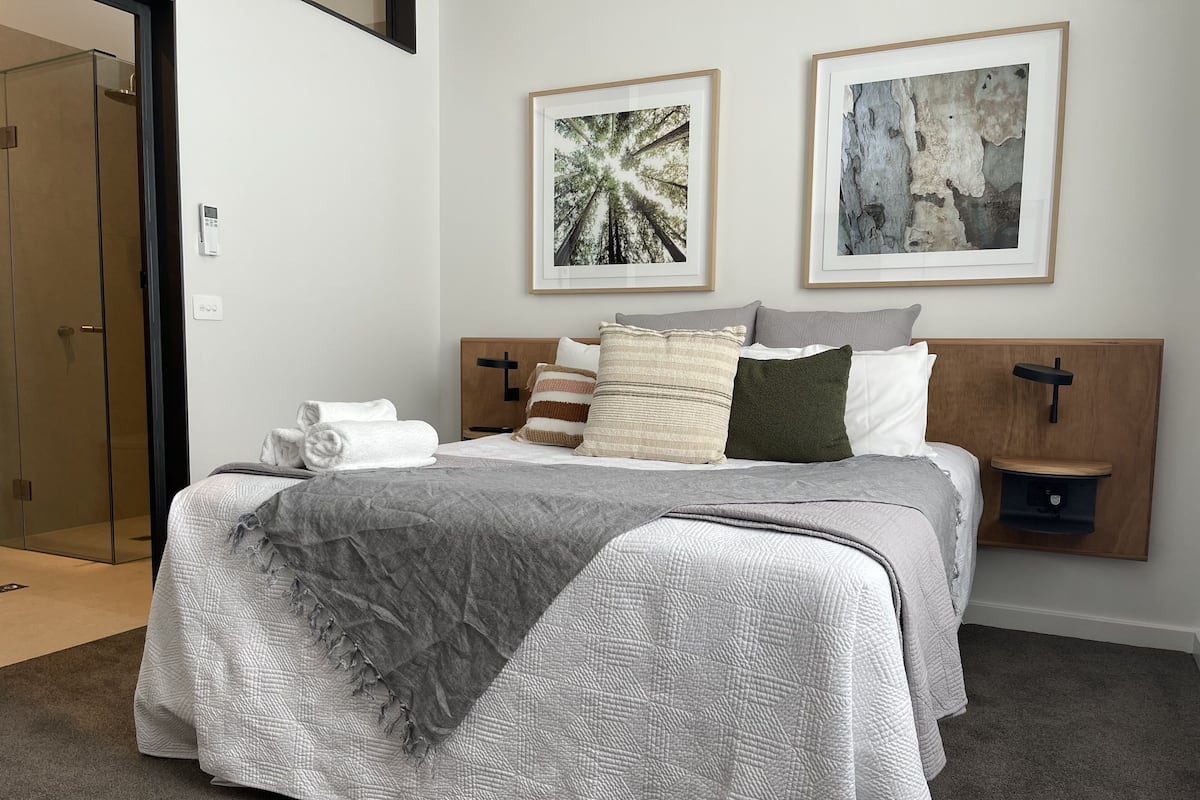 Una cama bien hecha con colcha gris, una toalla blanca y varios almohadones. Se observan dos cuadros enmarcados sobre la cama.