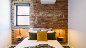 A bedroom with a brick wall, a window, a bed with pillows, and bedside tables with lamps.