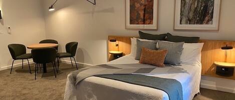 Un dormitorio moderno con una cama grande, una mesita de noche, un escritorio con sillas y dos cuadros enmarcados en la pared.