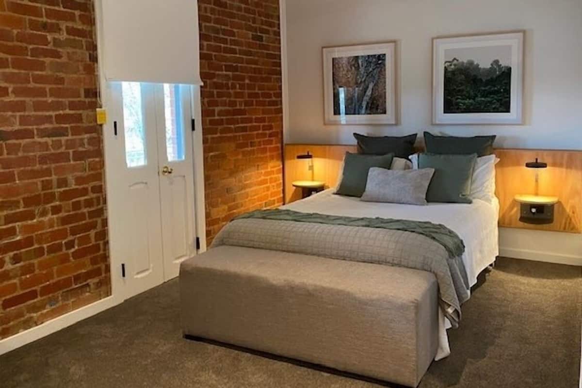 Un dormitorio con pared de ladrillo, una cama doble con almohadas y colcha, dos mesitas de noche con lámparas y dos cuadros enmarcados en la pared.