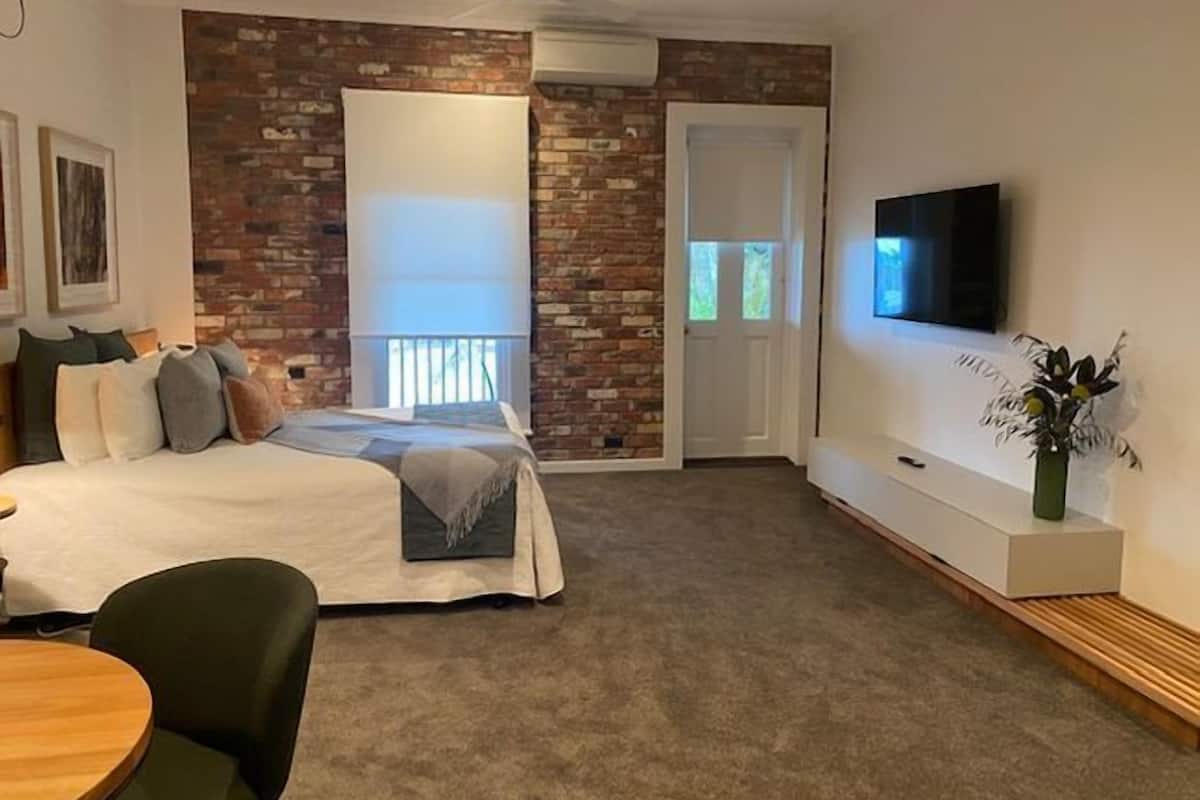Un dormitorio con pared de ladrillo decorativa, una cama con almohadas, un televisor en la pared y una pequeña mesa con sillas.