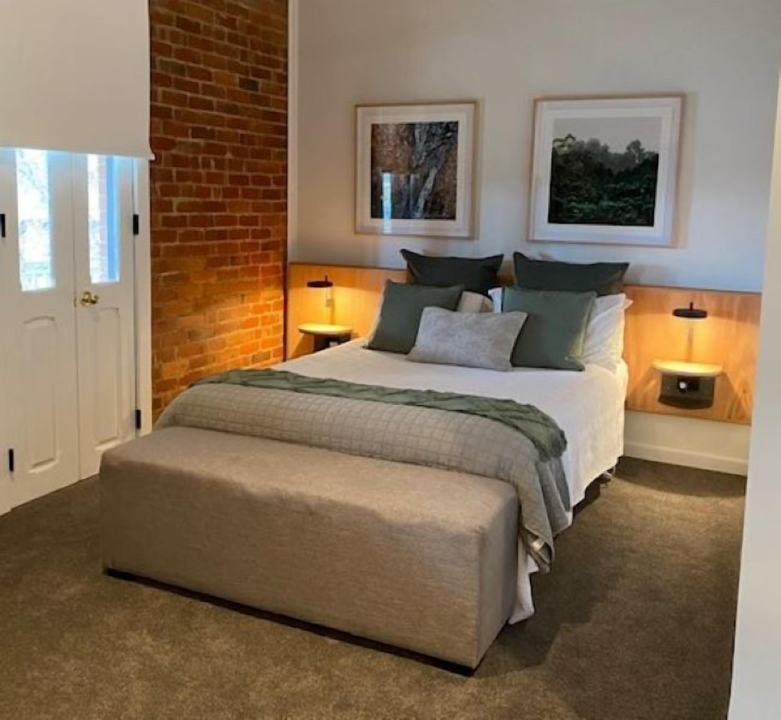 Un dormitorio con una cama, dos mesitas de noche, un banco, una pared de ladrillo y dos cuadros enmarcados.