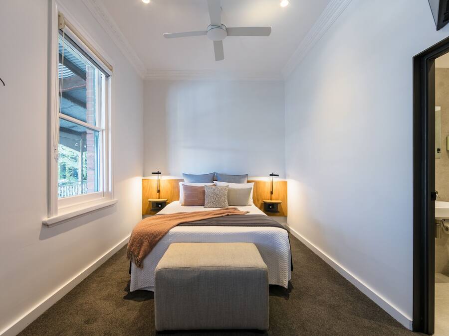 Quarto com cama, luminárias de cabeceira, ventilador de teto, janela e banheiro visível por uma porta aberta.