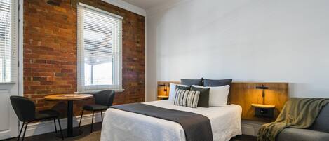 Habitación de hotel con pared de ladrillo, cama con cabecero de madera, una mesa redonda con sillas, sofá y mesita de noche con lámpara.