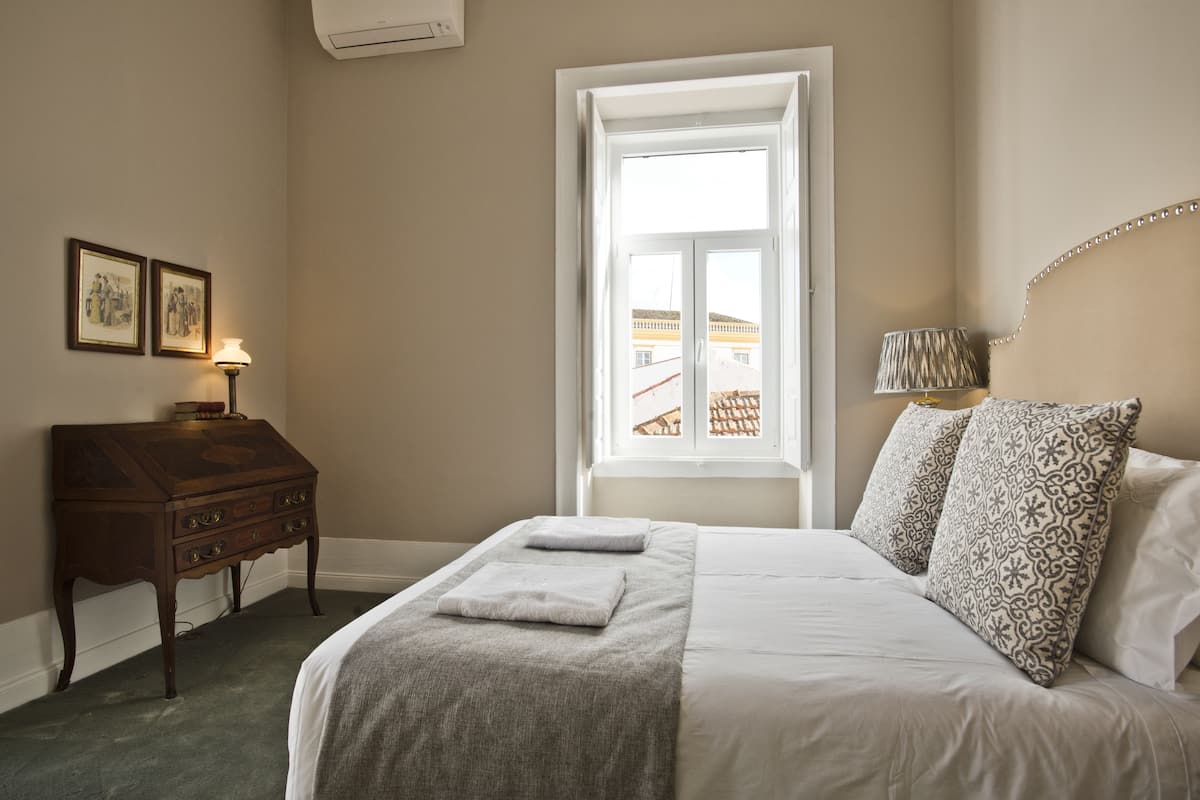 A neatly arranged bedroom with a large bed, a bedside table, a lamp, and a window with a view of buildings outside.