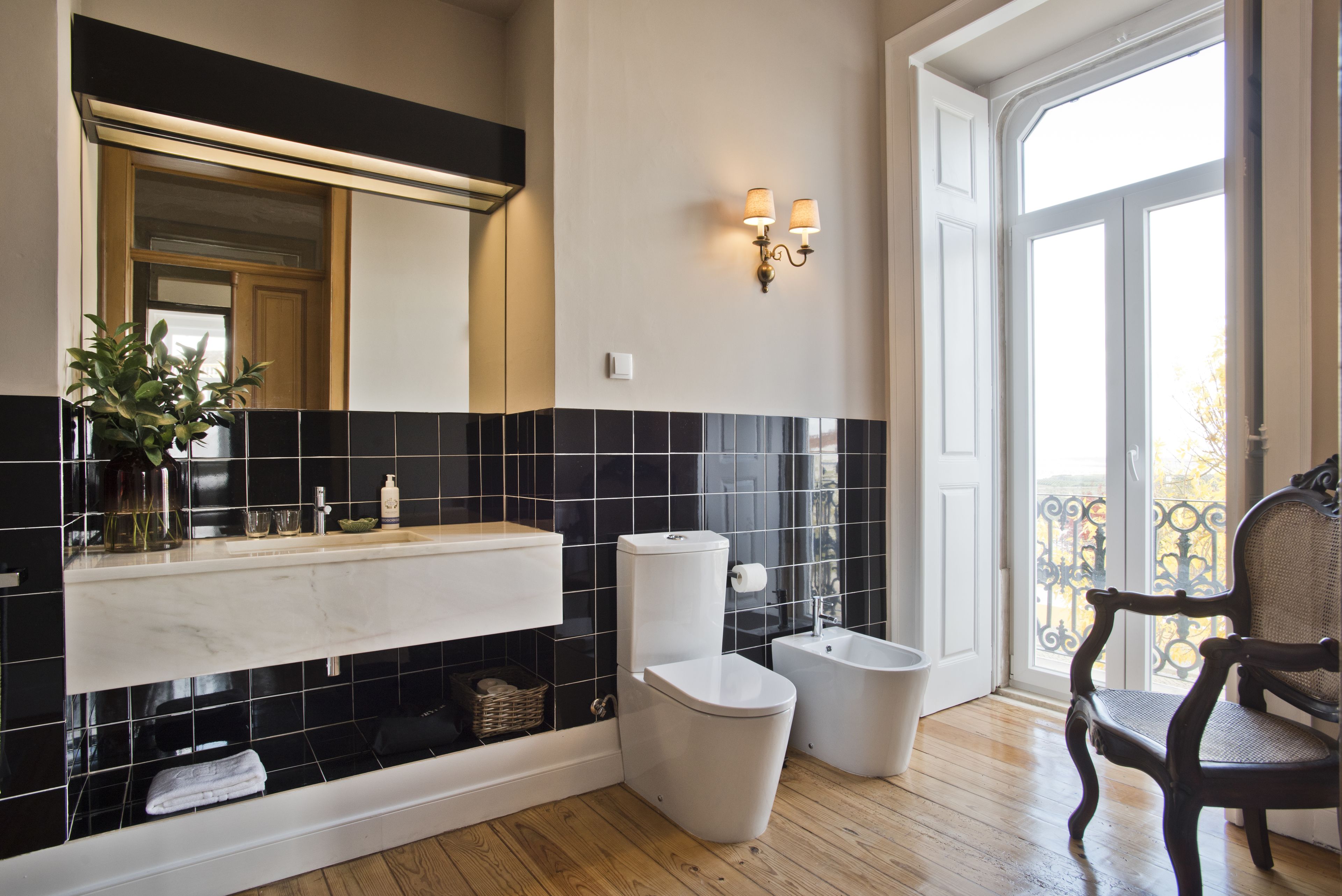 Une salle de bain avec un mur recouvert de carreaux noirs et blancs, un lavabo blanc, des toilettes et un bidet. Il y a un miroir au-dessus du lavabo et une chaise près de la fenêtre.