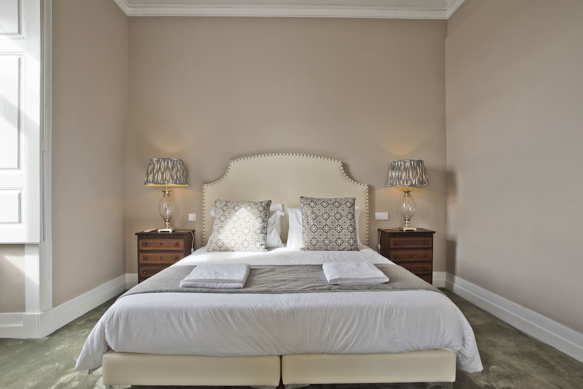 A neatly made bed with a headboard, two bedside tables with lamps, and a window on the left.