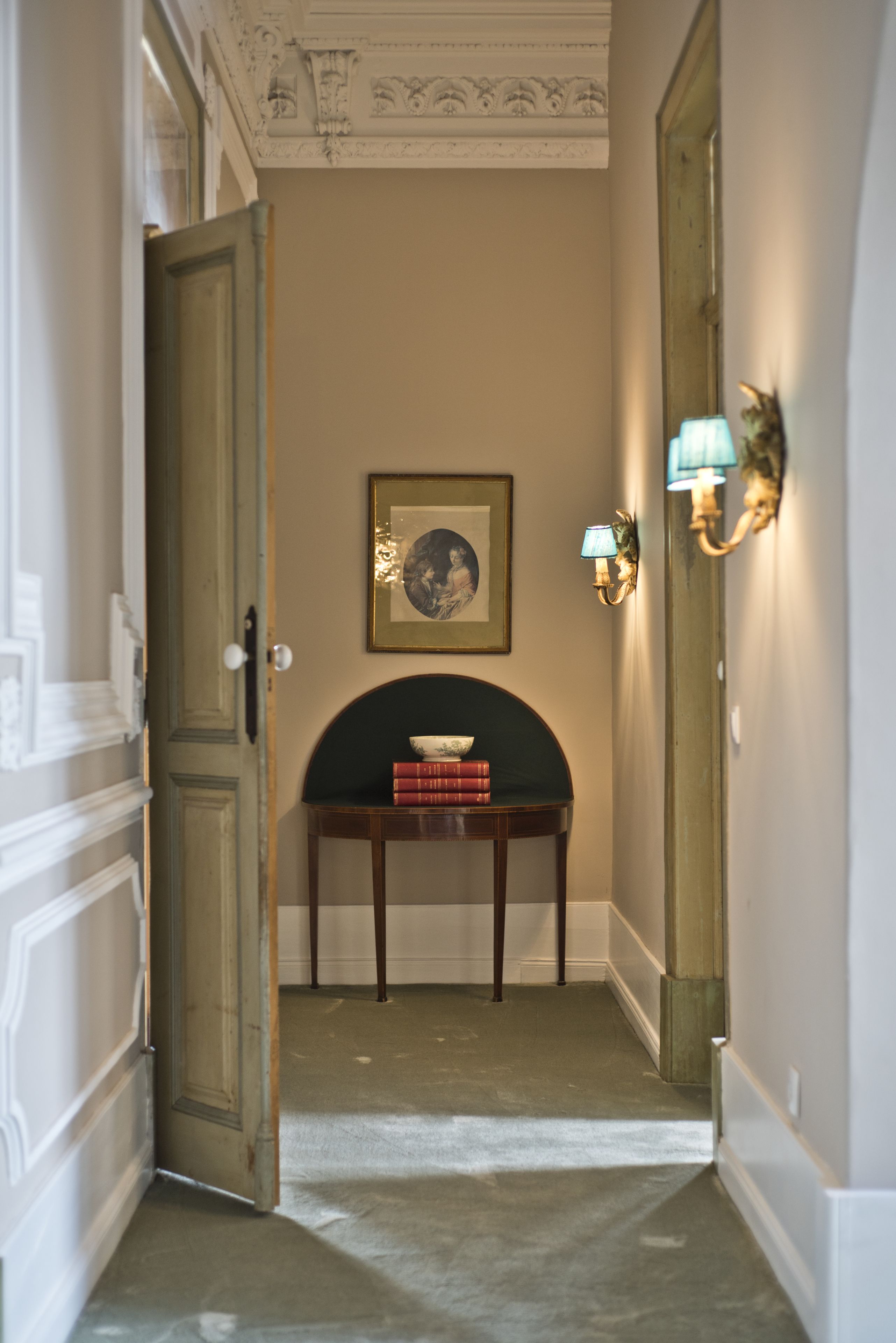 Un pasillo con una puerta, una lámpara de pared, un cuadro enmarcado y una pequeña mesa con libros.