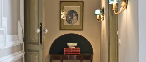 Un pasillo con una puerta, una lámpara de pared, un cuadro enmarcado y una pequeña mesa con libros.