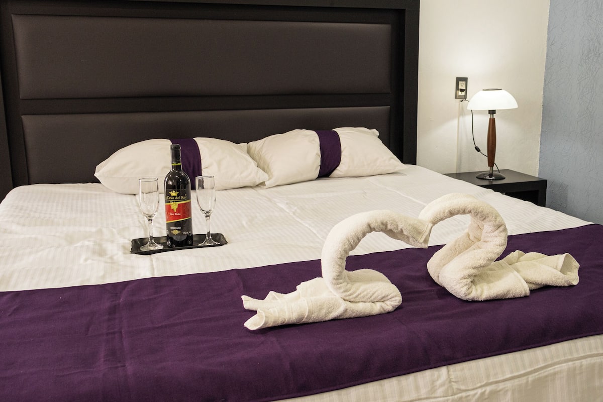 Un lit bien fait, recouvert d’une couverture violette, une bouteille de vin et deux verres à vin posés sur un plateau.