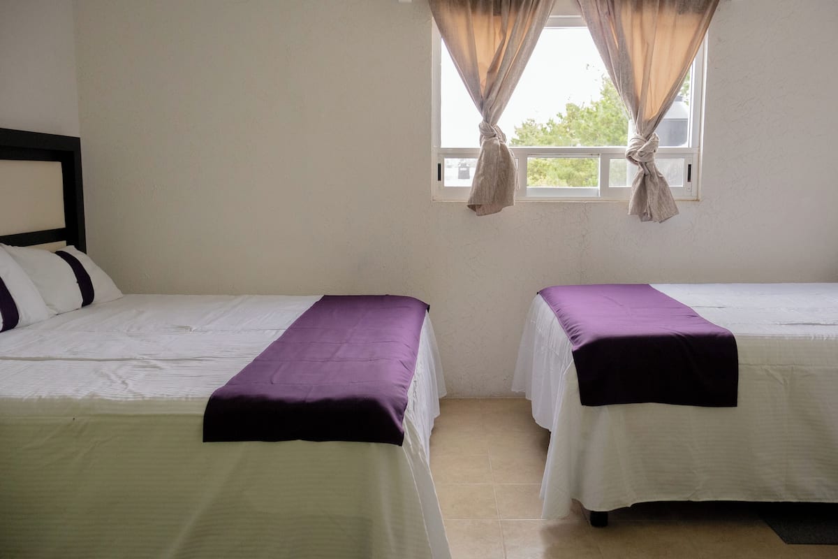 Une chambre avec deux lits, chacun recouvert d’une courtepointe blanche et d’une couverture violette. Il y a une fenêtre avec des rideaux attachés sur les côtés, et les murs sont de couleur unie.