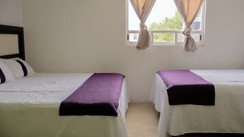 A room with two beds, each with a white bedspread and a purple blanket. There is a window with curtains tied back, and the walls are plain.