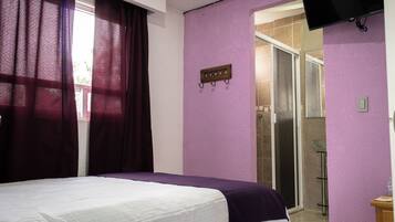 Habitación de hotel con cama, paredes moradas, zona de ducha y televisor montado.