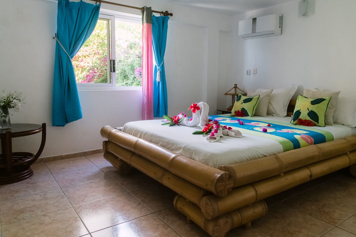 Una habitación con cama, ventana con cortinas, una mesa con un florero y aire acondicionado instalado en la pared.