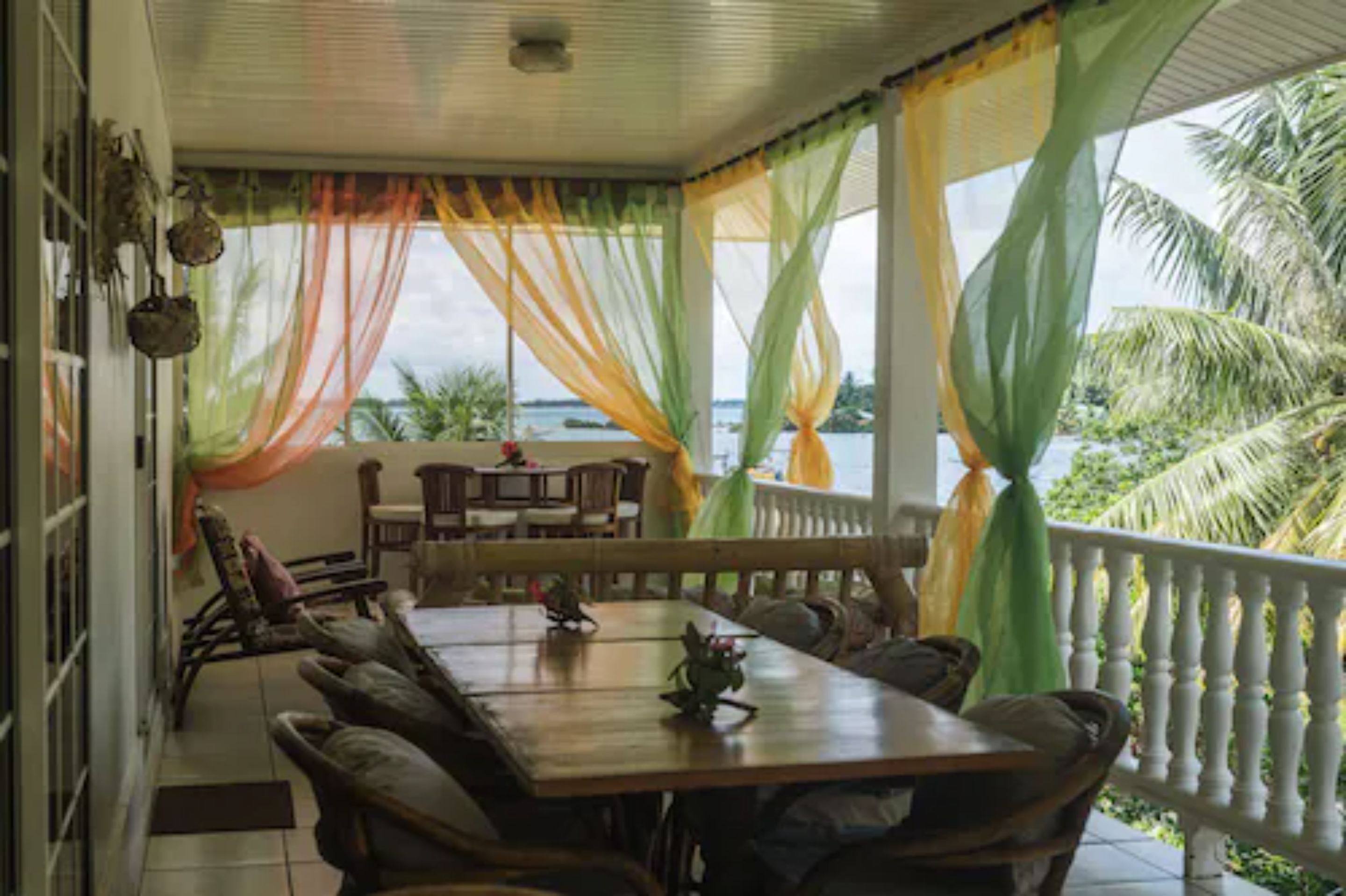 A covered outdoor dining area with a wooden table and chairs, draped with colorful curtains, overlooking a body of water and palm trees.