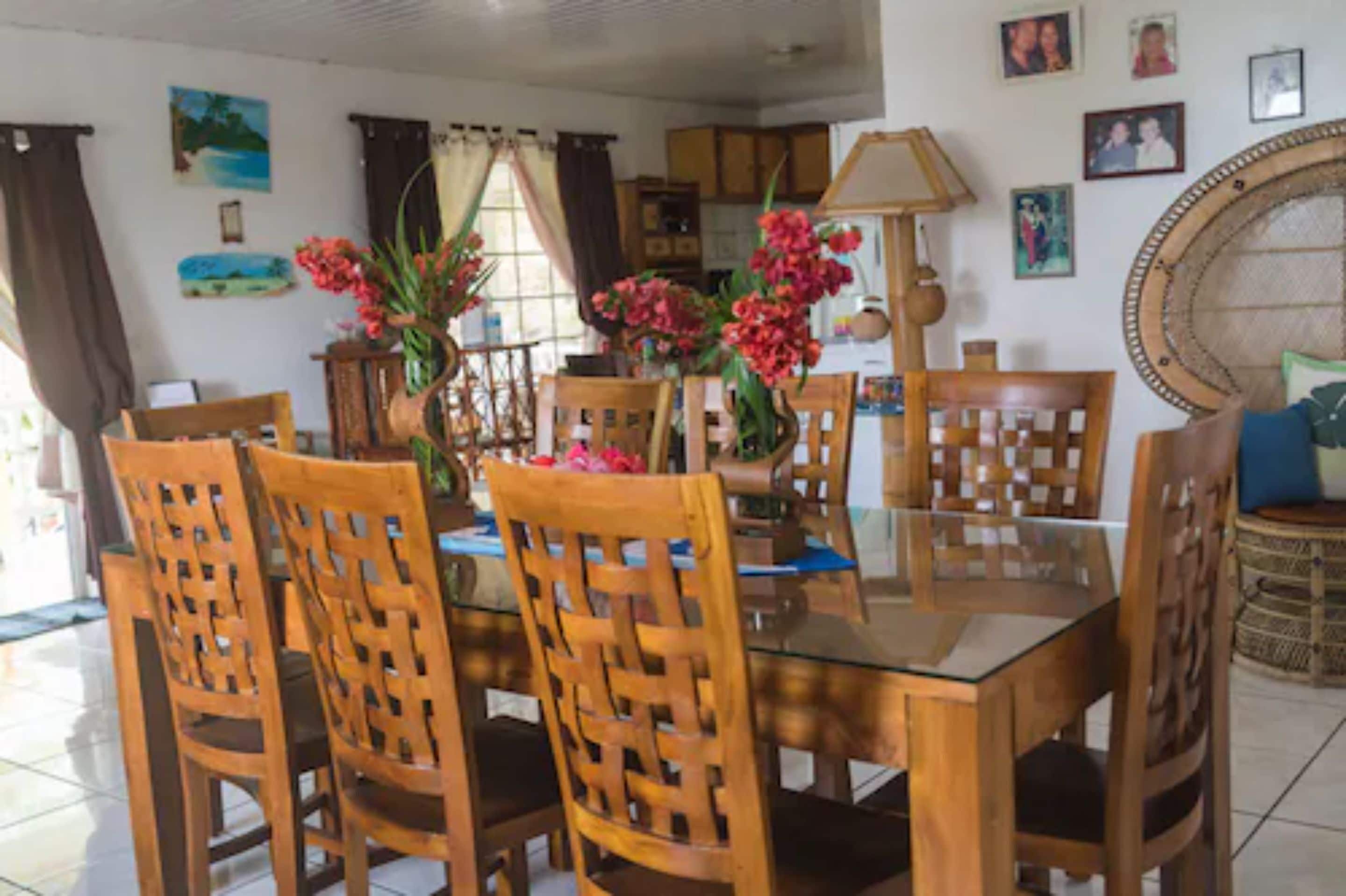 Un coin repas avec une table en verre et des chaises en bois, entouré de murs décorés d’images encadrées et agrémenté d’un grand fauteuil en osier.