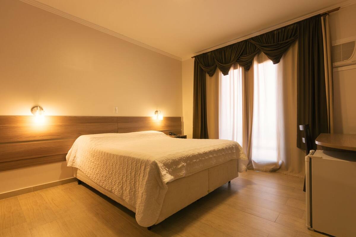 A hotel room with a bed, two wall-mounted lights, a wooden headboard, a window with curtains, and a small refrigerator.