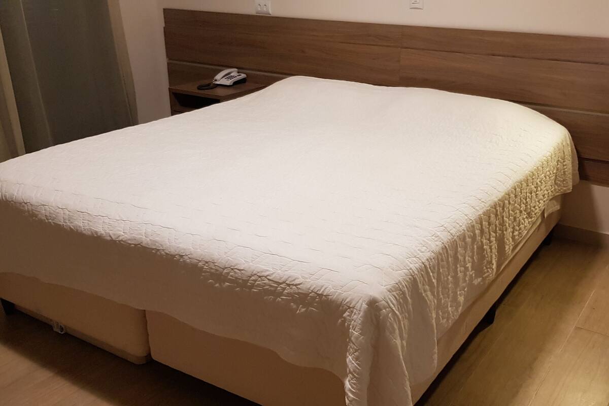 A neatly made bed with a wooden headboard, a bedside table with a phone, and a light on the wall.