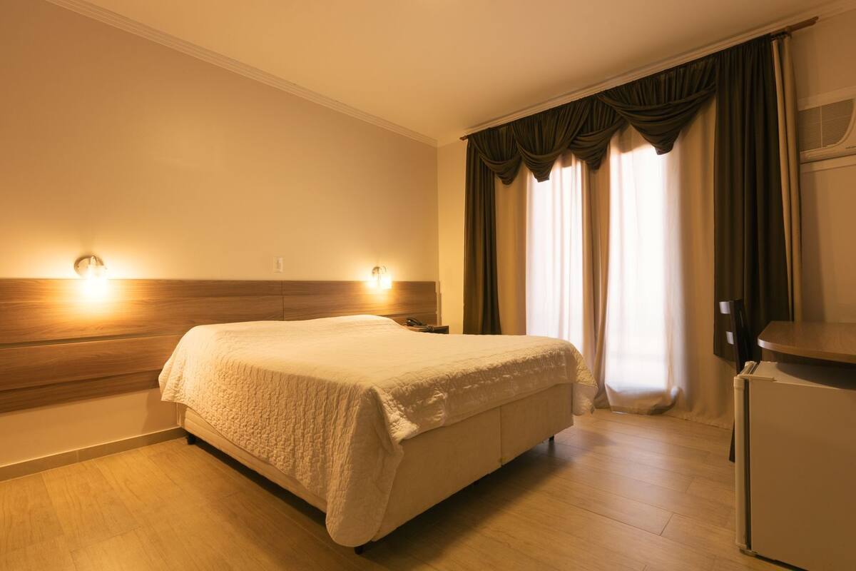 A hotel room with a bed, two wall-mounted lights, a wooden headboard, a window with curtains, and a refrigerator.